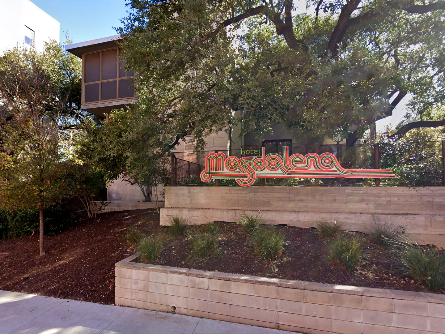 hotel-magdalena-hotel-magdalena-3-web copy Entrance to Hotel Magdalena on South Congress in Austin, Texas, with the neon script sign on a terraced concrete retaining wall and native grass planting beds along the sidewalk.