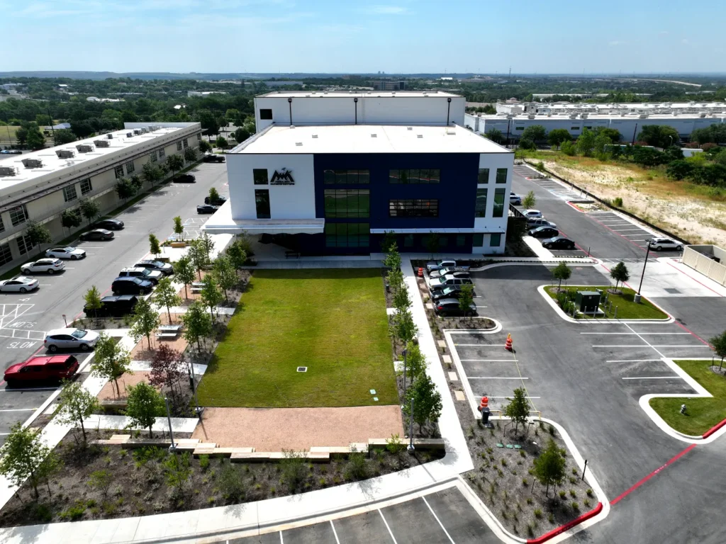 mesa-rim-austin-dji-0065-web Aerial view of the completed Mesa Rim Climbing Centers in Austin, Texas, showing the white and navy gym building fronted by a central lawn, tree-lined pedestrian plaza and parking on both sides, with site grading, drainage and paving by Mahoney Engineering.
